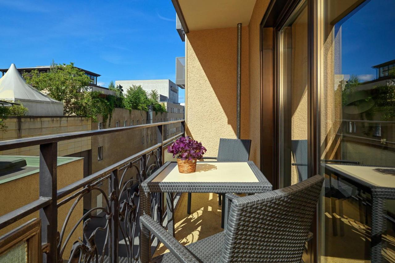 Отель The Westin Grand Берлин Экстерьер фото The photo depicts a balcony featuring a small table and two chairs. The table is set with a potted purple flower, adding a touch of decoration. The balcony has a wrought iron railing and glass walls, allowing for outdoor views. The background shows g