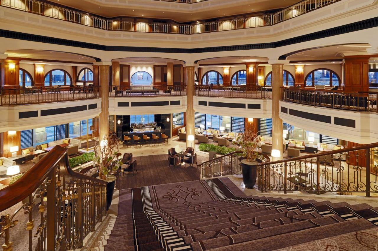 Отель The Westin Grand Берлин Экстерьер фото The photo depicts the interior of a luxurious hotel lobby. It features a grand multi-level design with a sweeping staircase that leads down to a spacious area. The lobby has elegant architectural elements, including large arched windows that allow na