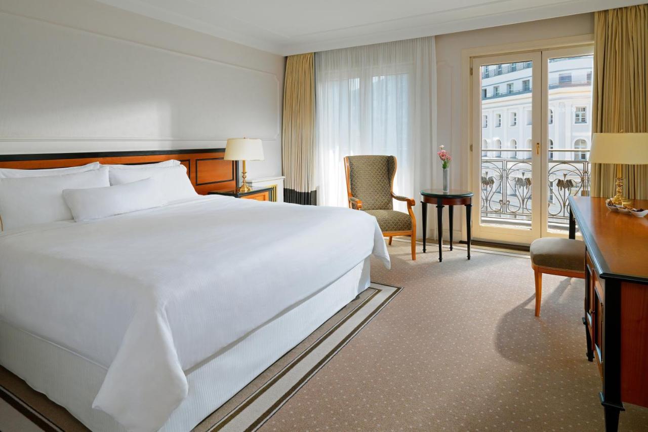 Отель The Westin Grand Берлин Экстерьер фото The photo shows a hotel room featuring a large bed with white linens, a bedside lamp, and a small armchair. There is a table next to the chair, and large windows with sheer curtains allow natural light to fill the space. The decor is simple and elega