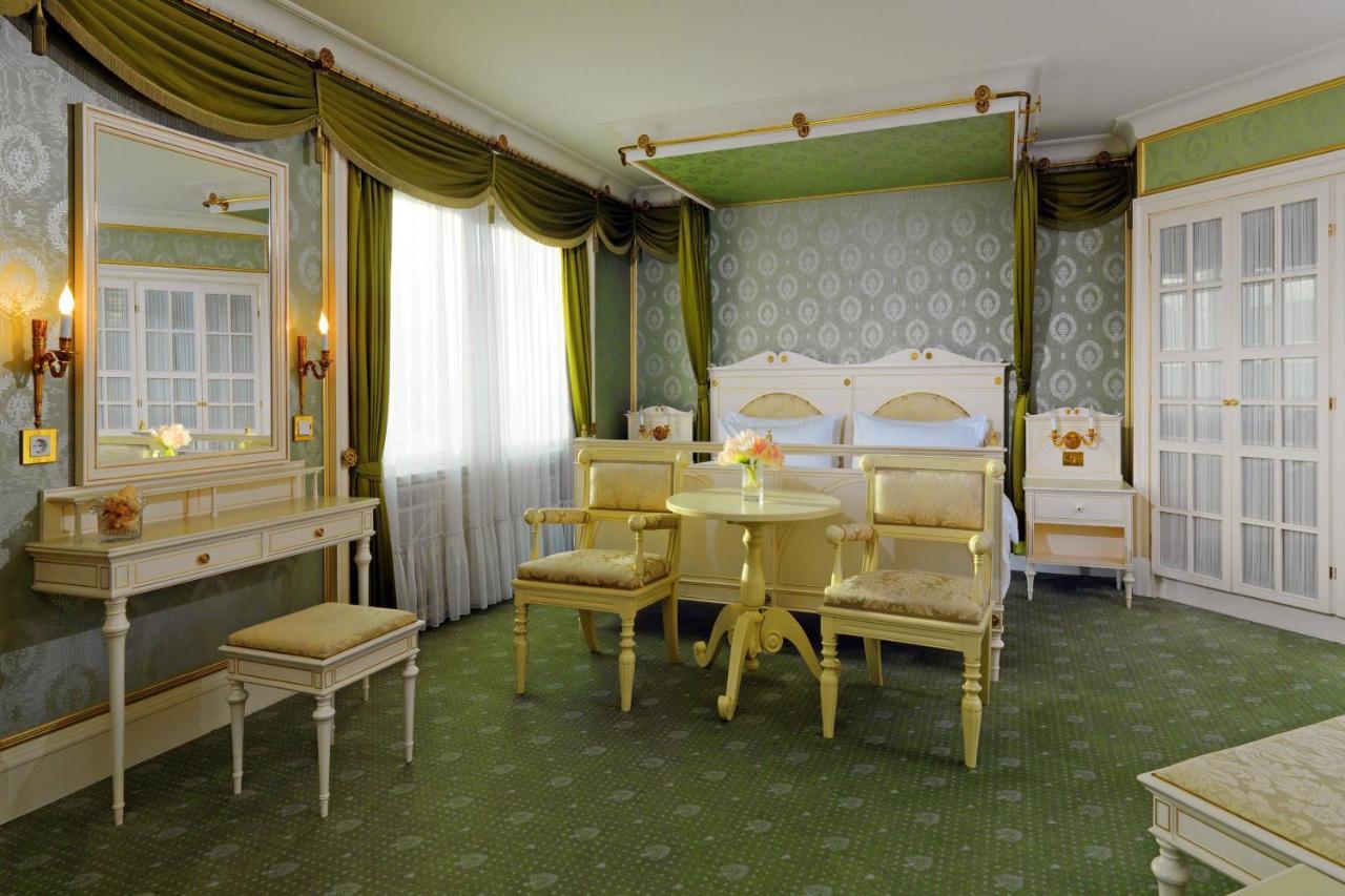 Отель The Westin Grand Берлин Экстерьер фото The photo shows a beautifully decorated hotel room with a classic and elegant design. The room features a large bed with an ornate headboard, flanked by bedside tables. There are two upholstered chairs and a small round table in the center, providing