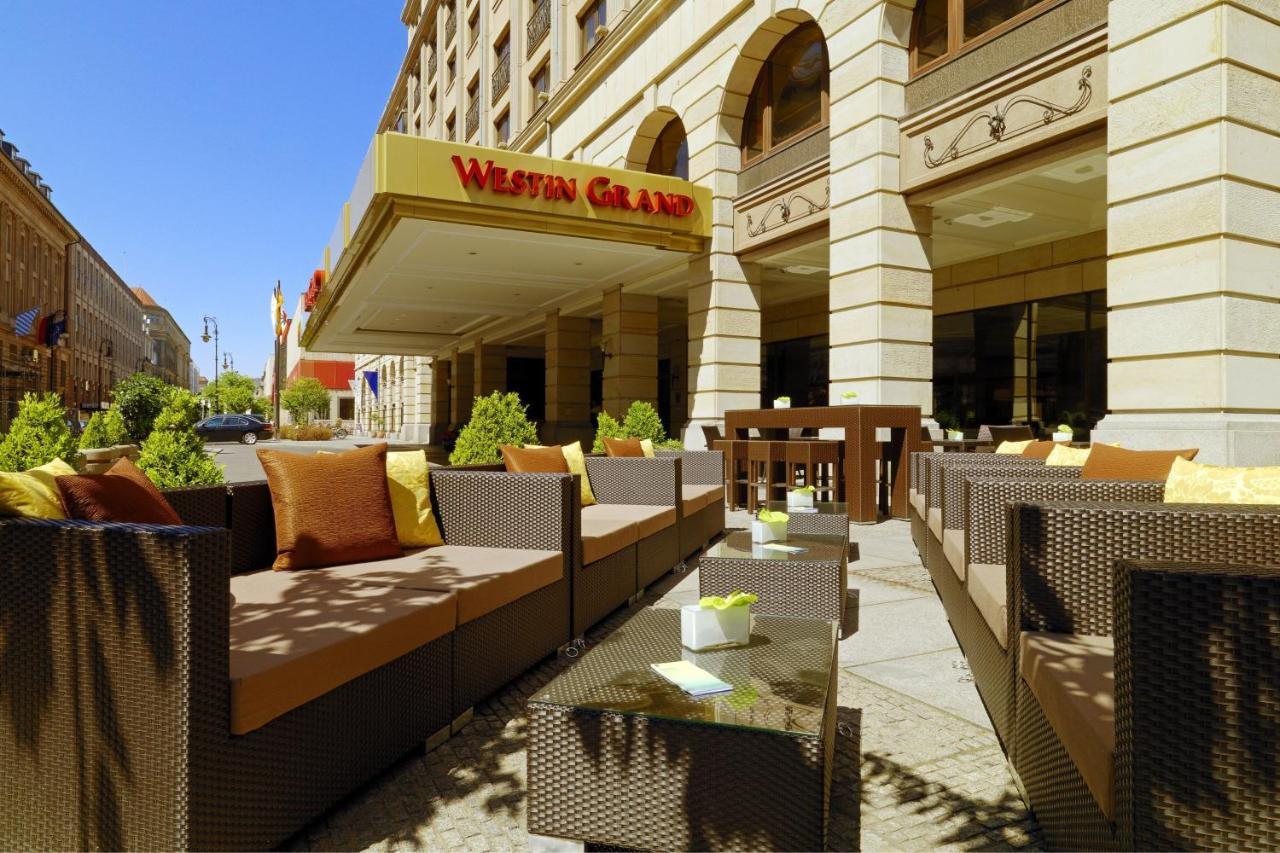 Отель The Westin Grand Берлин Экстерьер фото The photo shows an exterior view of a hotel with a prominent sign that reads "Westin Grand." In the foreground, there is a stylish outdoor seating area featuring brown wicker furniture, including couches and coffee tables. The seating is accented wit