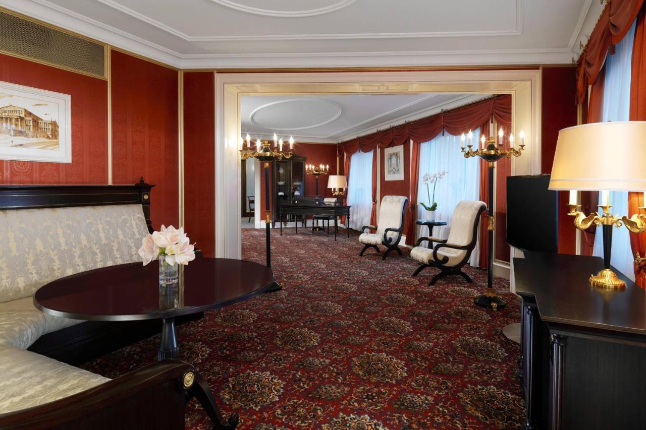 Отель The Westin Grand Берлин Экстерьер фото The photo shows an elegantly decorated hotel suite or living space. The interior features rich red wallpaper and a plush patterned carpet, creating a warm and inviting atmosphere. There's a round table with a vase of flowers in the foreground, and tw