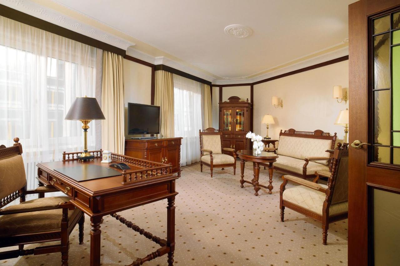 Отель The Westin Grand Берлин Экстерьер фото The photo shows an elegant room with a classic design. It features a seating area with upholstered chairs and a sofa, all arranged around a coffee table. A wooden desk with a lamp is positioned nearby, and there is a television on a cabinet. Soft cur