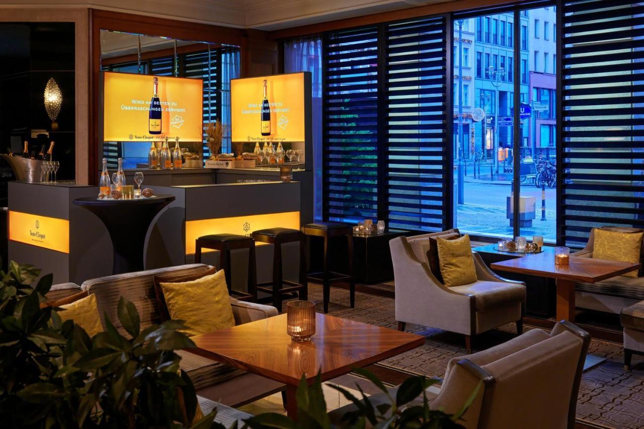 Отель The Westin Grand Берлин Экстерьер фото The photo shows a modern bar or lounge area with a contemporary design. There are several tables and comfortable seating arranged around the space, featuring cozy chairs and light-colored cushions. The bar area is well-lit, with stylish displays illu