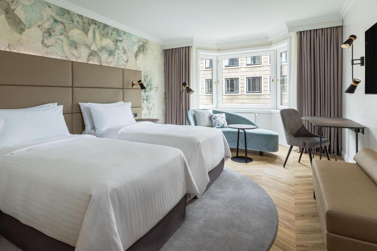 Отель The Westin Grand Берлин Экстерьер фото The photo shows a modern hotel room featuring two twin beds with white linens arranged neatly. The walls are adorned with a subtle floral wallpaper, creating a serene atmosphere. There's a small seating area near a large window, which allows for natu