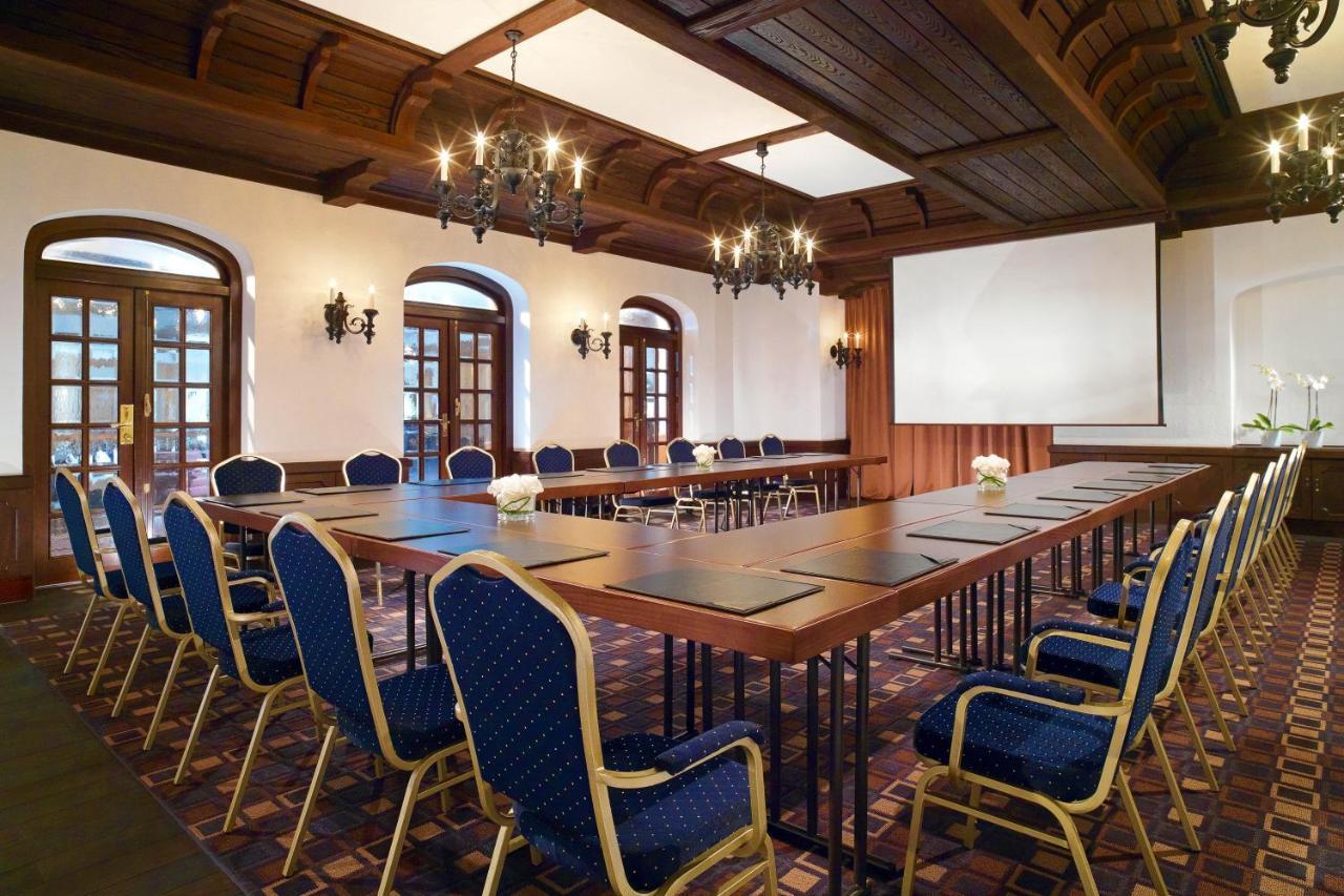Отель The Westin Grand Берлин Экстерьер фото The photo shows a conference room with an elegant design. There is a long rectangular table set up in the center, surrounded by upholstered chairs with blue fabric. The room features wooden ceiling beams and decorative lighting fixtures, including ch