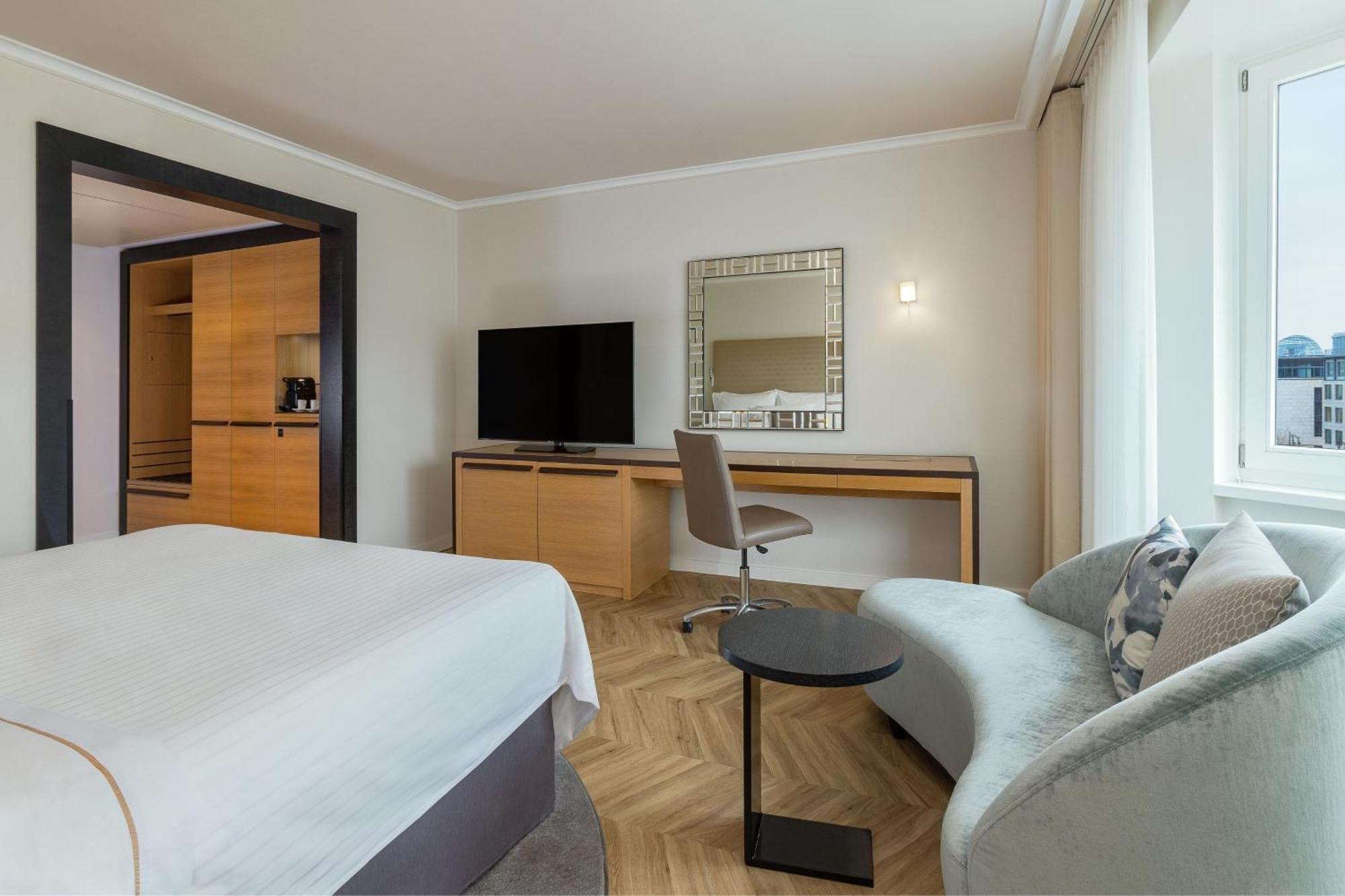 Отель The Westin Grand Берлин Экстерьер фото The photo shows a modern hotel room that features several elements:

1. **Bed**: A neatly made bed with a light-colored duvet and white sheets.
2. **Furniture**: There is a small round table next to a contemporary gray sofa, and a desk with a chair p