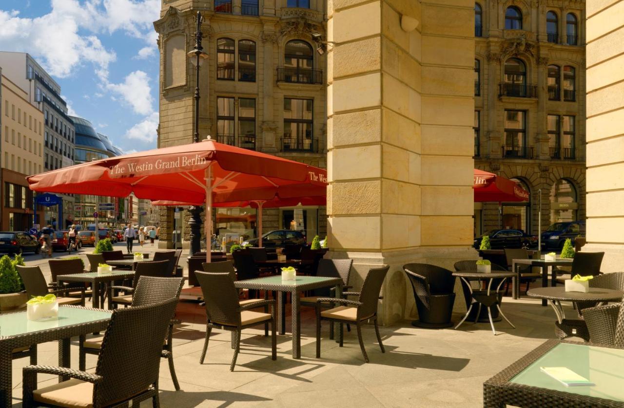 Отель The Westin Grand Берлин Экстерьер фото The photo shows an outdoor café area with several tables and chairs arranged under large red umbrellas. There are several potted plants on the tables, adding a touch of greenery. The café is situated in an urban setting, with impressive buildings in 